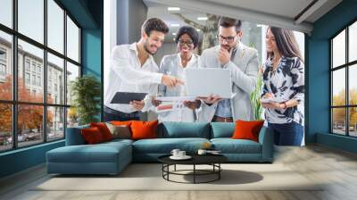 Multiethnic group of inspired young people standing and discussing new project on laptop in office building. Wall mural