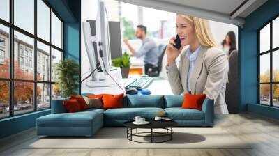 Smiling business woman working on computer and talking on mobile phone in office Wall mural