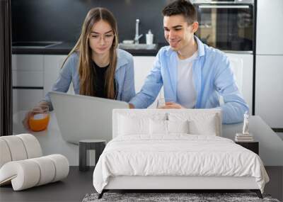 Smiling teenage boy holding glass with juice and using laptop accompanied with his girl friend at home. Wall mural