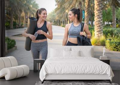 Sporty women going to park for fitness workout. Two beautiful fitness female friends in sportswear holding gym bag, yoga mat and water bottle in park. Wall mural