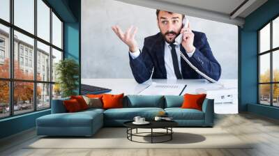 Successful young entrepreneur in suit sitting at work desk, talking on the phone and explaining something. Wall mural