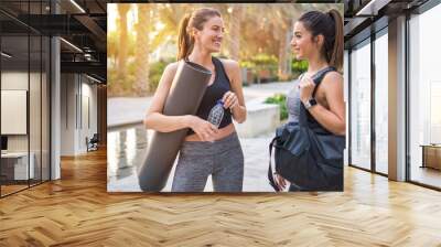 Two beautiful fitness female friends in sportswear holding gym bag, yoga mat and water bottle talking in the park. Sporty girl ready for training. Wall mural