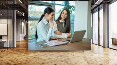 Two brunette business women, talking, advising each other while using laptop in the office. Wall mural