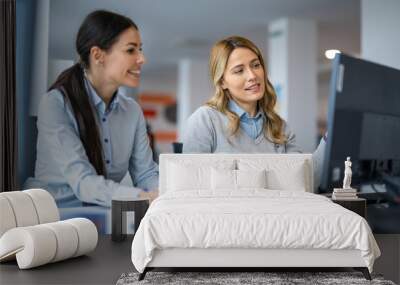 Two female business colleagues working together and discussing project on computer while sitting at desk in office. Wall mural