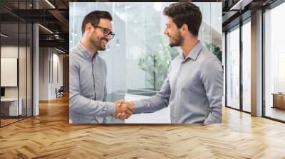 Two smiling businessmen shaking hands together while standing in modern office. Wall mural