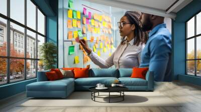 Young mixer race businesswoman manager explaining strategy ideas on sticky notes on glass wall to male African American colleague looking at strategy scrum presentation. Business project planning. Wall mural