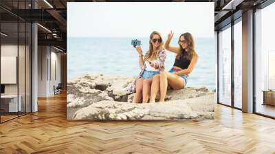 Two girlfriends on a sea shore taking selfies and sitting on rocks Wall mural