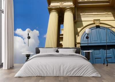 Close-up of classical architectural columns and arched doorway against a bright blue sky with fluffy clouds, showcasing historical elegance. Wall mural