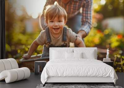 Happy little boy having fun in a wheelbarrow pushing by dad in domestic garden on warm sunny day. Wall mural