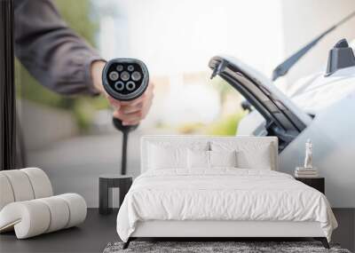 Side view, close-up shot of unrecognizable Asian woman holding type 2 EV charger plug, standing in front of an electrical car hood. Wall mural