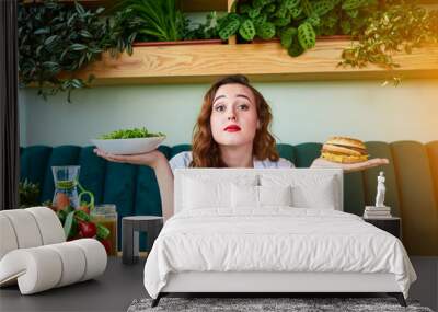 Beautiful young woman decides eating hamburger or fresh salad in kitchen. Cheap junk food vs healthy diet Wall mural