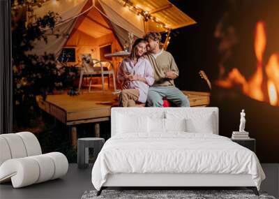 Happy couple relaxing in glamping on summer evening and drinking wine near cozy bonfire. Luxury camping tent for outdoor recreation and recreation. Lifestyle concept Wall mural