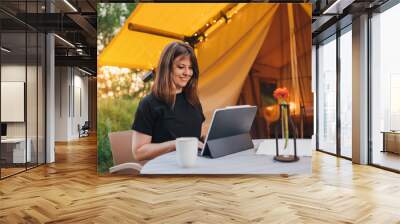 Smiling Woman freelancer using a laptop on a cozy glamping tent in a sunny day. Luxury camping tent for outdoor summer holiday and vacation. Lifestyle concept Wall mural