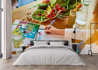 Woman dietitian in medical uniform with tape measure working on a diet plan sitting with different healthy food ingredients in the green office on background. Weight loss and right nutrition concept Wall mural