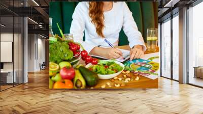 Woman dietitian in medical uniform with tape measure working on a diet plan sitting with different healthy food ingredients in the green office on background. Weight loss and right nutrition concept Wall mural