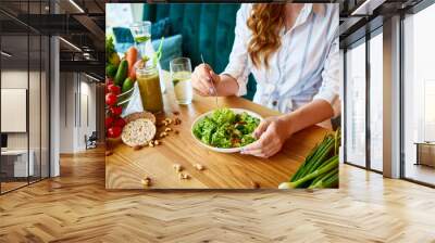 Young happy woman eating salad in the beautiful interior with green flowers on the background and fresh ingredients on the table. Healthy food concept Wall mural