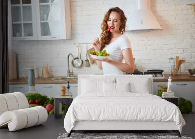 Young happy woman eating salad in the beautiful kitchen with green fresh ingredients indoors. Healthy food concept Wall mural