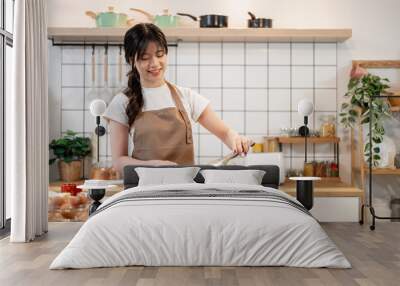 A beautiful, happy Asian woman in an apron is enjoying cooking breakfast in the kitchen. Wall mural