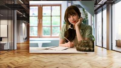 A beautiful dreamy Asian girl is writing her diary while her cute cat is on a table Wall mural