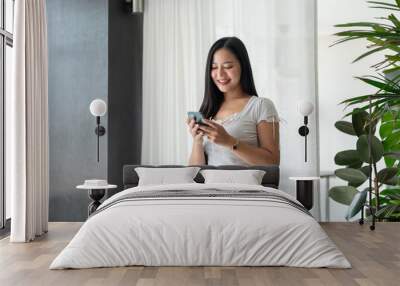 A charming Asian woman is smiling at text messages on her phone while standing by the window. Wall mural