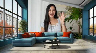 A cheerful Asian woman is standing by a window in a room, smiling and waving her hand at the camera. Wall mural