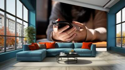 A cropped image of a woman using her smartphone at a table by the window on a sunny day. Wall mural