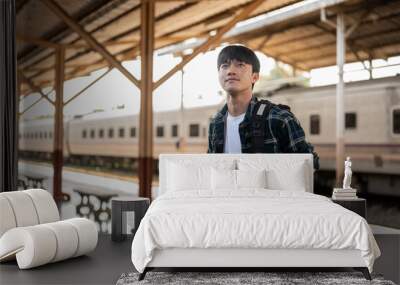 A handsome, happy young Asian man tourist backpacker standing at a platform in a railway station. Wall mural