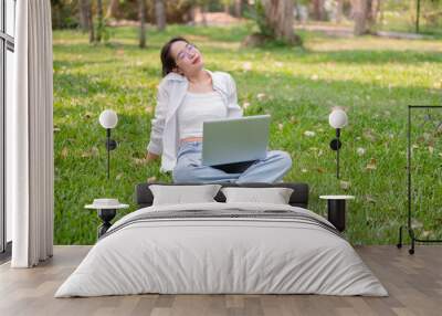 A happy, relaxed Asian woman sits on the grass in a park with her laptop, enjoying the sunlight. Wall mural