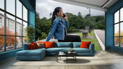 A positive Asian woman in a denim jacket is talking on the phone with someone on a bench in a park. Wall mural