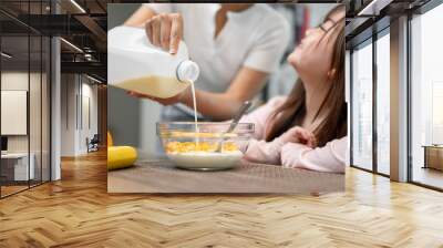 Close-up of a caring mom pouring milk from a gallon into a cereal bowl for her daughter's breakfast. Wall mural