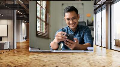 Handsome and happy Asian businessman using his mobile phone at his desk. Wall mural