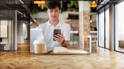 Handsome Asian man is focused on reading chat on his phone while working remotely from a coffee shop Wall mural