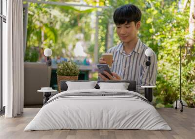 Handsome Asian man is having a coffee and using his smartphone while sitting at a table in a garden. Wall mural