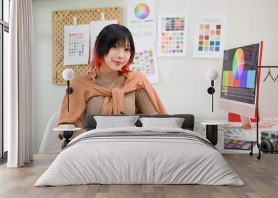 Smiling Asian female graphic designer or interior designer sits at her desk in her studio. Wall mural