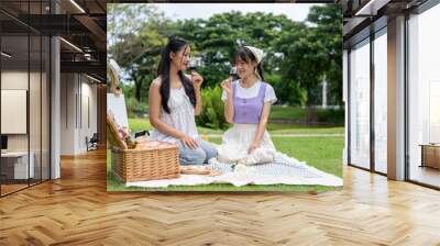 Two happy, charming young Asian women are enjoying red wine together during a picnic in a green park Wall mural