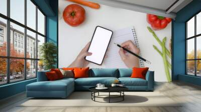 Young woman noting diet plan while looking information on mock-up smartphone on table with fresh vegetables Wall mural