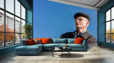 Aged senior man in cap and glasses sitting on bench against blue sky Wall mural