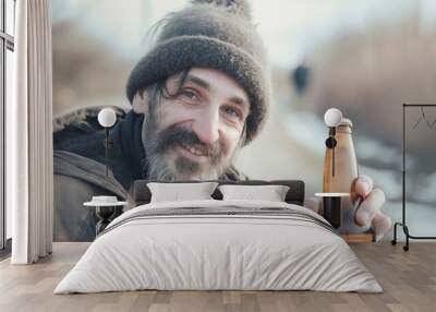 Bearded man with beanie holding bottle of beer, smiling cheerfully. Outdoor winter setting with blurred background, wearing warm jacket and casual attire Wall mural