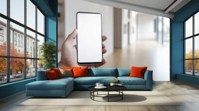 Caucasian hand holding a modern black smartphone with a blank screen in a bright modern office space Wall mural