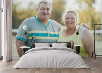 Happy middle aged couple standing on tennis court holding rackets and smiling at camera. They appear active and enjoying game together highlighting their bond and enthusiasm for sports Wall mural