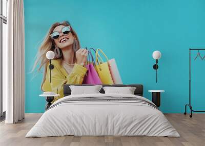 Happy woman with long blonde hair wearing sunglasses and large earrings, holding colorful shopping bags against a bright blue background Wall mural