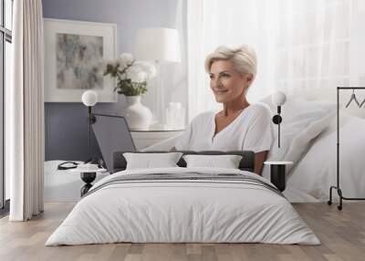 Smiling senior woman using laptop while sitting comfortably on bed in bright bedroom with soft lighting and elegant decor Wall mural