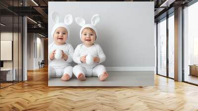 Two Adorable Twin Babies Dressed in White Onesies and Bunny Ears Holding White Eggs and Smiling Happily at the Camera Wall mural