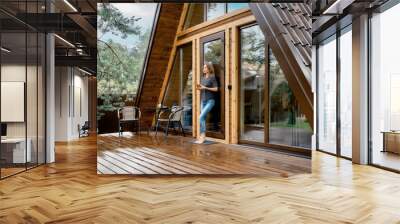 Pensive woman stands on the terrace of a wooden bungalow with a glass of herbal tea Wall mural