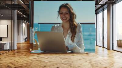 A female wearing white blouse is smiling and typing on a laptop on a seaside terrace Wall mural