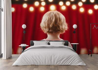 Rear view of a child standing on stage with bright lights and red curtains, ready to perform in a theater production or school play. Wall mural