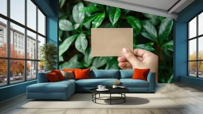 Hand holding blank brown paper card in nature background. Mockup of paper card note on green leaves background Wall mural