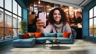 Portrait of cool cheerful girl having video-call with friend holding smart phone in hand shooting selfie on front camera with a cafe in the background.  Wall mural