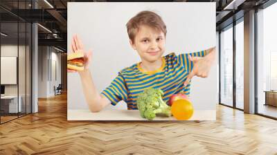 Young funny boy at the table refuses hamburger in favor of fruit and vegetables on a white background Wall mural