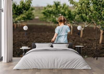 Little anonymous girl with two braids, walking on a dirt ground between peach trees in a garden. Wall mural